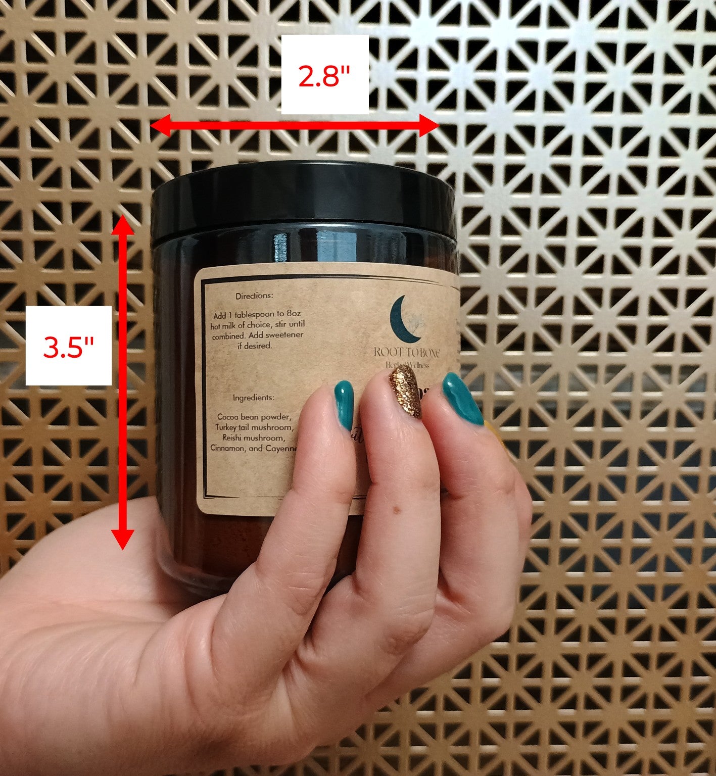 A person holding a small mug-sized container with a label that has text and a moon symbol. The container is brown with a black lid, and the person's hand is against a geometric pattern background.