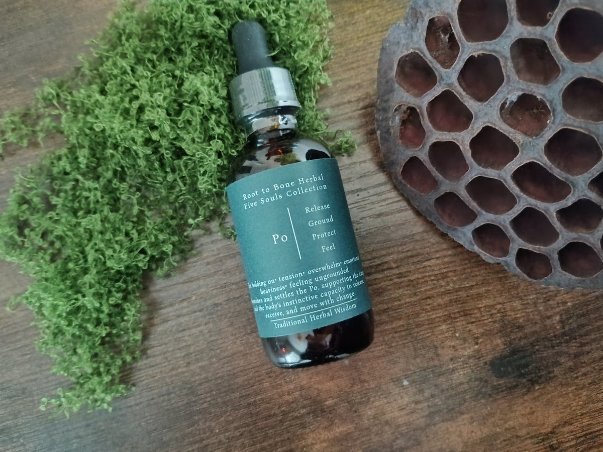 A small amber glass dropper bottle with a dark teal label sits on a wooden surface, styled with green moss on the left and a brown honeycomb pod on the right. The label shows the brand 'Root to Bone Herbal' and product name 'Po'