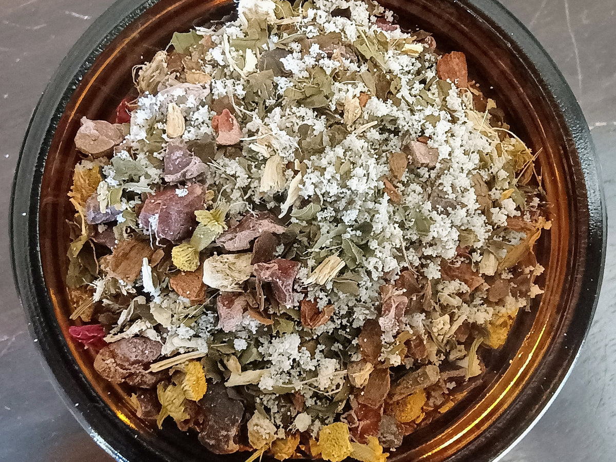 A top-down view of a dried herbal tea blend inside a dark brown ceramic bowl, featuring dried leaves, flowers, and white coconut milk crystals on a gray stone surface.