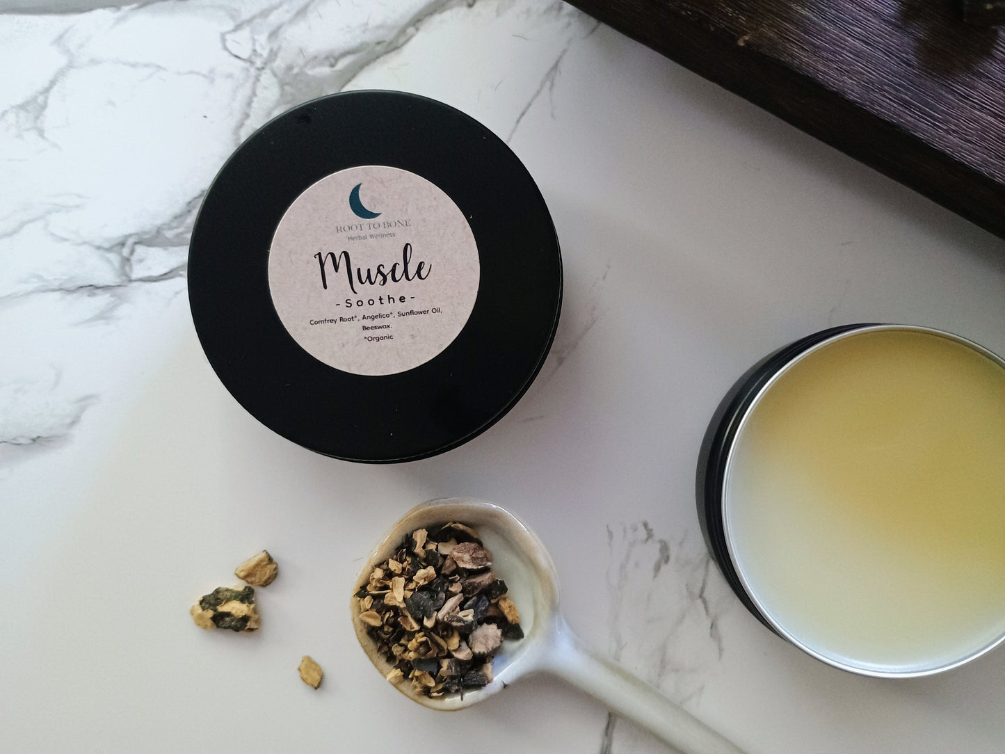 Black container with a white label, a spoon with dried herbs, and a open salve on a marble surface.