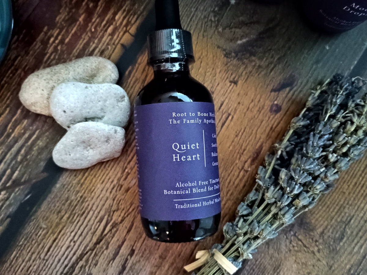 A dark amber glass dropper bottle with a purple label reading 'Quiet Heart' by Root to Bone and The Family Apothecary, styled on a rustic wooden surface with three smooth stones and a bundle of dried lavender.