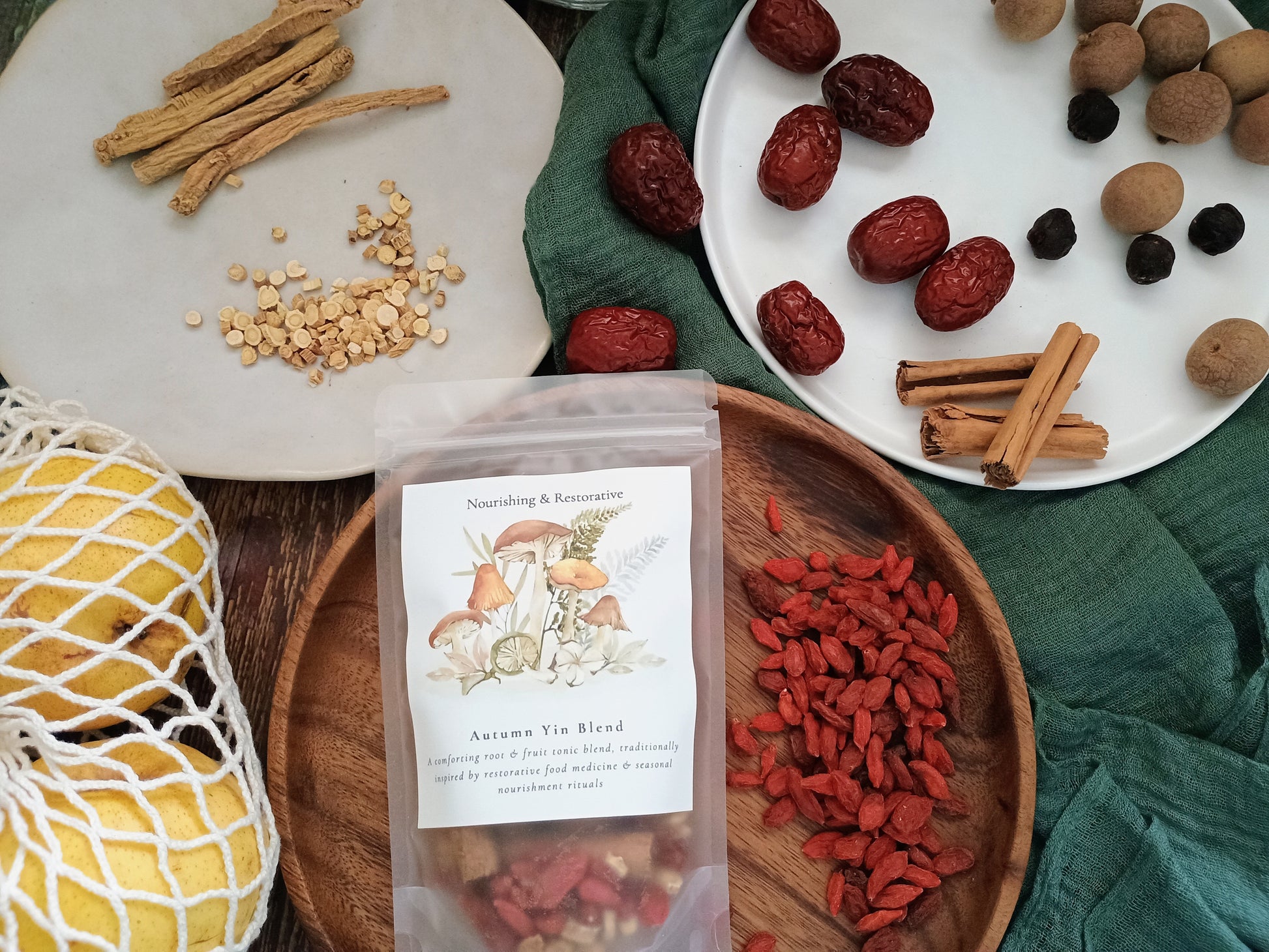 A stand-up pouch labeled 'Autumn Yin Blend' sits on a wooden tray, surrounded by loose herbal ingredients including red goji berries, cinnamon sticks, dried red dates, and tan seeds on plates and fabric.