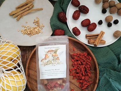 A stand-up pouch labeled 'Autumn Yin Blend' sits on a wooden tray, surrounded by loose herbal ingredients including red goji berries, cinnamon sticks, dried red dates, and tan seeds on plates and fabric.