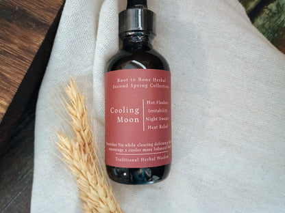 A dark amber glass dropper bottle with a black cap and a dusty rose label reading 'Cooling Moon' by Root to Bone Herbal, styled on a textured white linen cloth with dried wheat stalks.