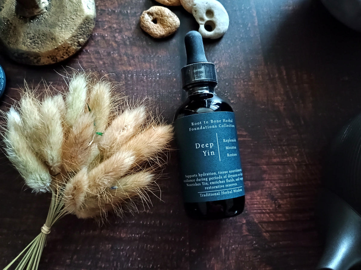 A dark amber glass dropper bottle labeled 'Deep Yin' by Root to Bone Herbs, placed on a dark wooden surface next to dried bunny tail grass and small decorative stones.