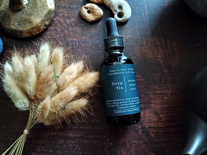 A dark amber glass dropper bottle labeled 'Deep Yin' by Root to Bone Herbs, placed on a dark wooden surface next to dried bunny tail grass and small decorative stones.