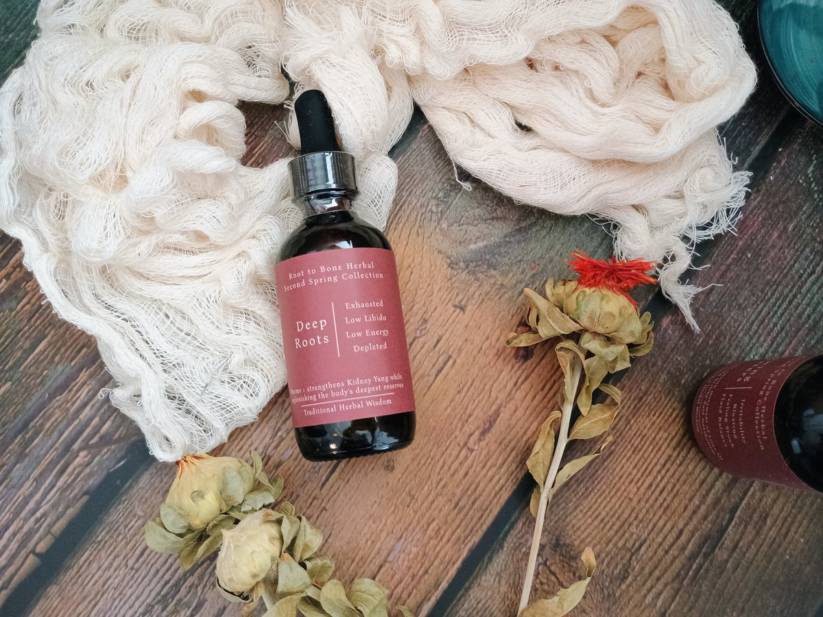 A dark amber glass dropper bottle labeled 'Deep Roots' by Root to Root Herbs, styled on a rustic wooden surface with cream gauze, dried flowers, and another partial bottle.