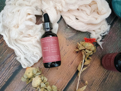 A dark amber glass dropper bottle labeled 'Deep Roots' by Root to Root Herbs, styled on a rustic wooden surface with cream gauze, dried flowers, and another partial bottle.