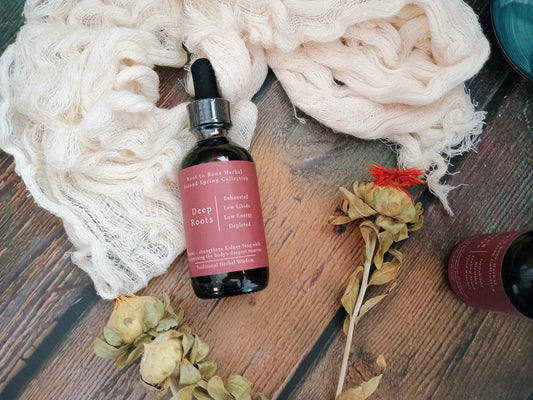 A dark amber glass dropper bottle labeled 'Deep Roots' by Root to Root Herbs, styled on a rustic wooden surface with cream gauze, dried flowers, and another partial bottle.