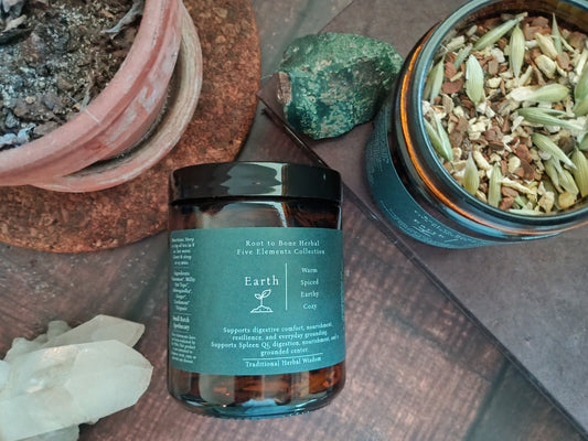 A jar of Root to Bone Herbal Earth Element tea 'Five Elements Collection' Spiced Infused Cozy herbal tea, styled on a wooden surface with crystals, a potted plant, and a tin of loose herbal ingredients.
