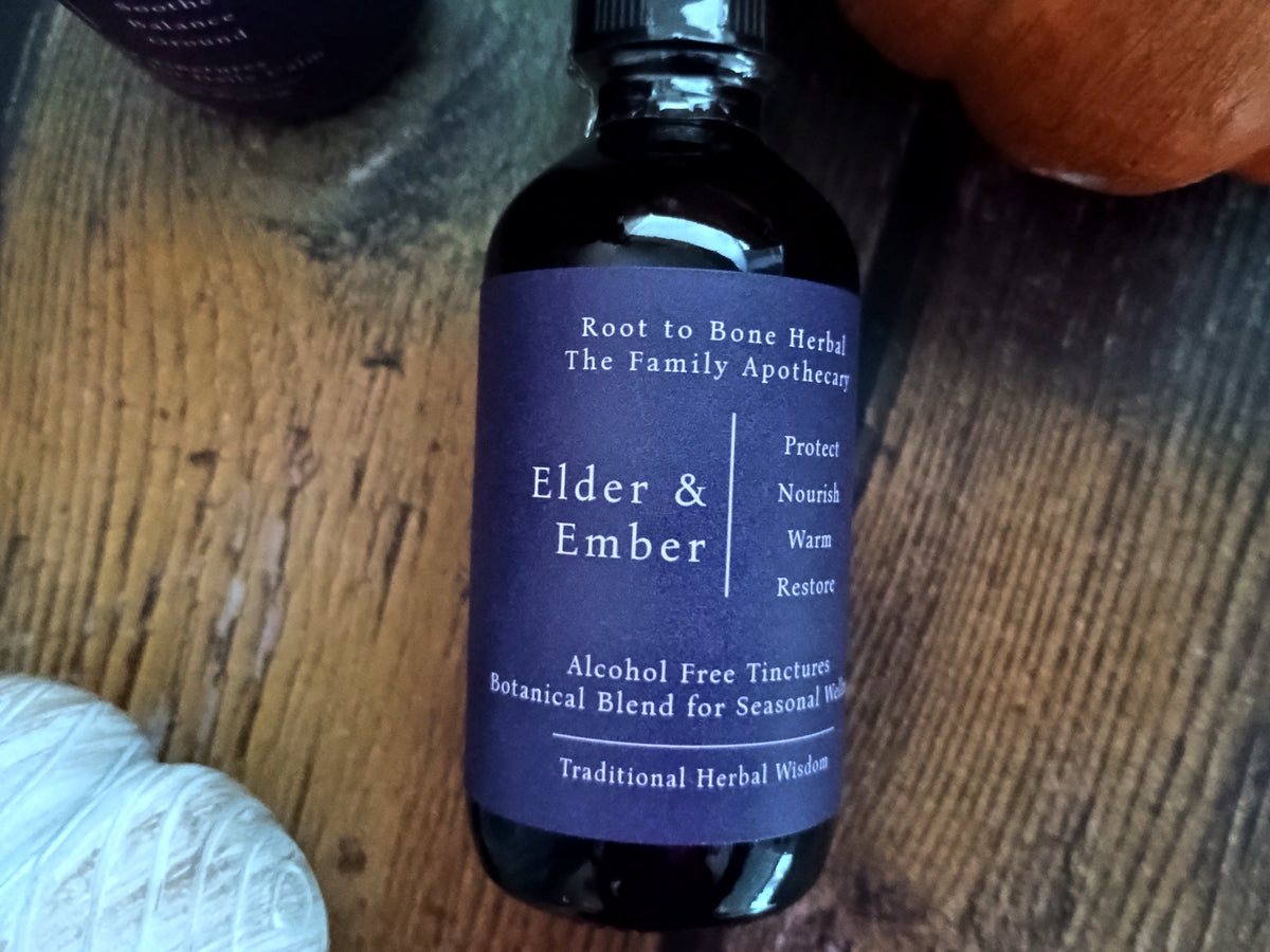 A dark amber glass bottle of Root to Bone Herbal 'Elder & Ember' tincture with a dark purple label, resting on a rustic wooden surface.
