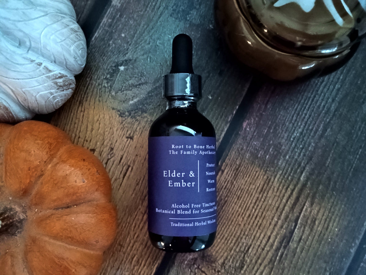 A dark amber glass dropper bottle labeled 'Elder & Ember' by Root to Bone Herbal, styled on a rustic dark wood surface with a small orange pumpkin, a white decorative object, and a brown glass bowl nearby.