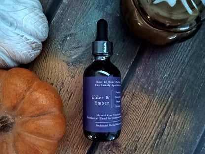A dark amber glass dropper bottle labeled 'Elder & Ember' by Root to Bone Herbal, styled on a rustic dark wood surface with a small orange pumpkin, a white decorative object, and a brown glass bowl nearby.
