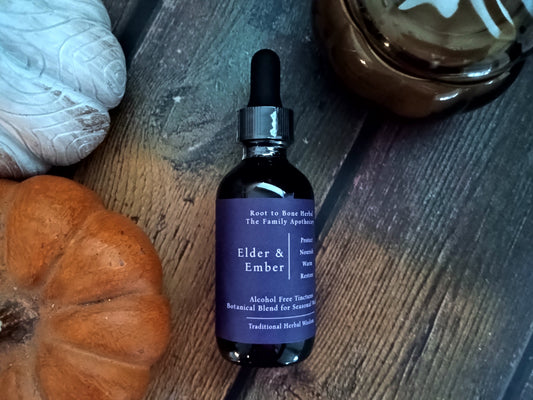 A dark amber glass dropper bottle labeled 'Elder & Ember' by Root to Bone Herbal, styled on a rustic dark wood surface with a small orange pumpkin, a white decorative object, and a brown glass bowl nearby.