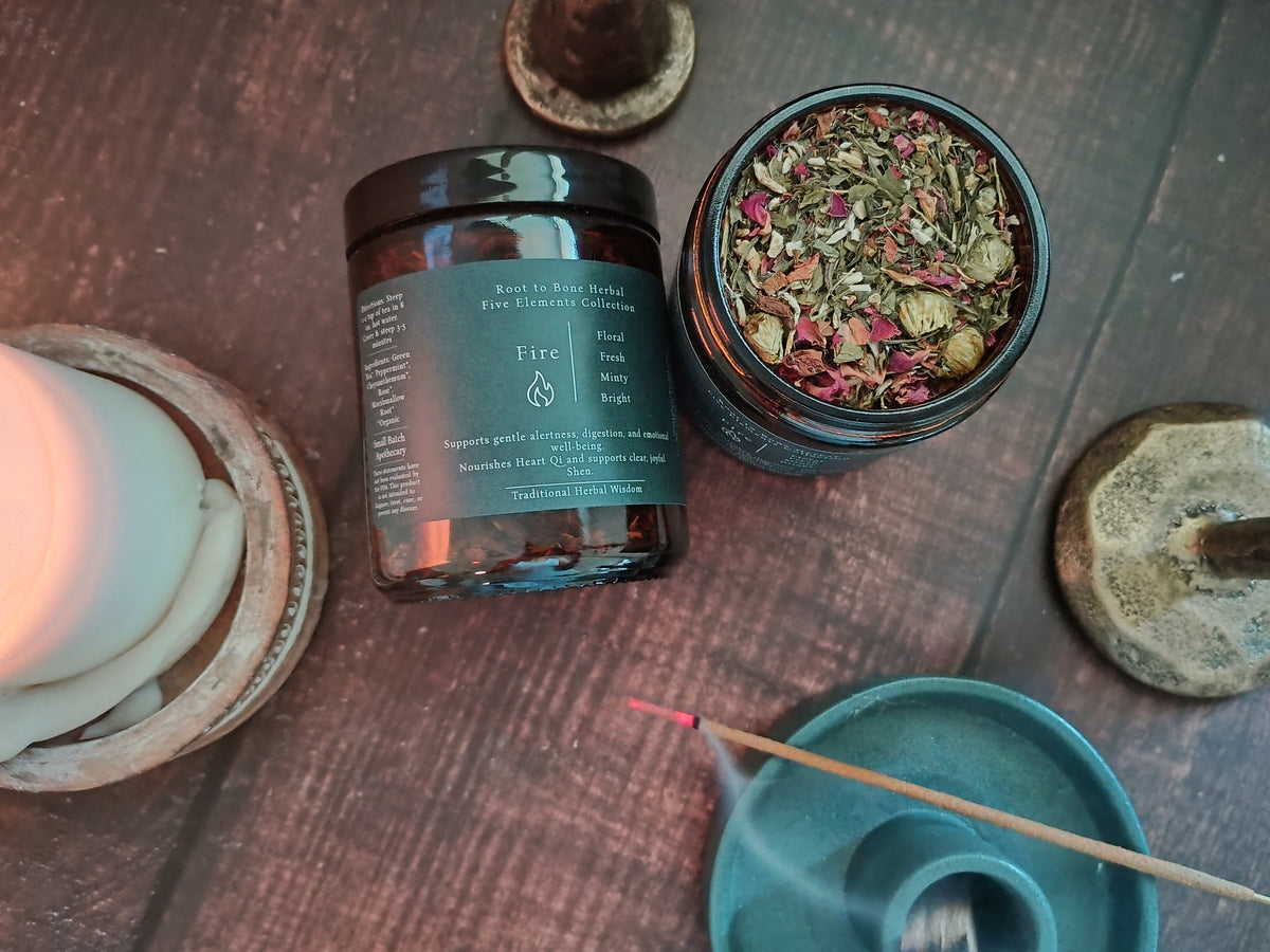An amber glass jar candle labeled 'Fire' by Root to Bone Herbs, shown next to an open jar displaying dried rose petals and herbs. The setup is styled on a dark wooden surface with a lit white candle, a teal incense holder, and other rustic spiritual props.