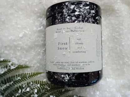 A dark amber glass jar candle with a white label reading 'Root to Bone Herbal Trees Collection First Snow' and 'soft creamy', placed on a bed of white ice crystals with a green fern leaf at the bottom left.