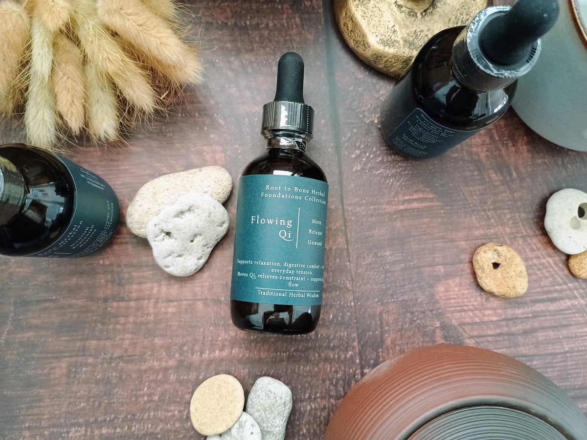 A dark amber glass dropper bottle labeled 'Flowing Qi' with a teal label and black dropper cap, styled on a wooden surface with stones and dried grass.