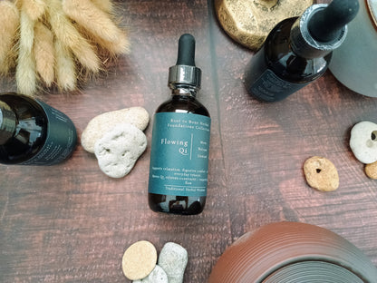 A dark amber glass dropper bottle labeled 'Flowing Qi' with a teal label and black dropper cap, styled on a wooden surface with stones and dried grass.