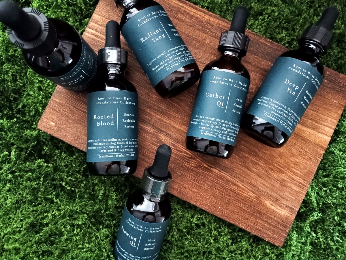 Six dark amber glass dropper bottles with teal labels arranged on a wooden cutting board over bright green grass. The labels read 'Roots in Bone Herbal Foundations Collective' and include names such as Radiant Yang, Gather Qi, Deep Yin, and Rooted Blood.