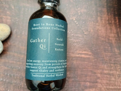 An amber glass bottle of Root to Bone Herbal Foundations Collection Gather Qi herbal tincture, featuring a dark teal label with white text, set on a dark wooden surface.