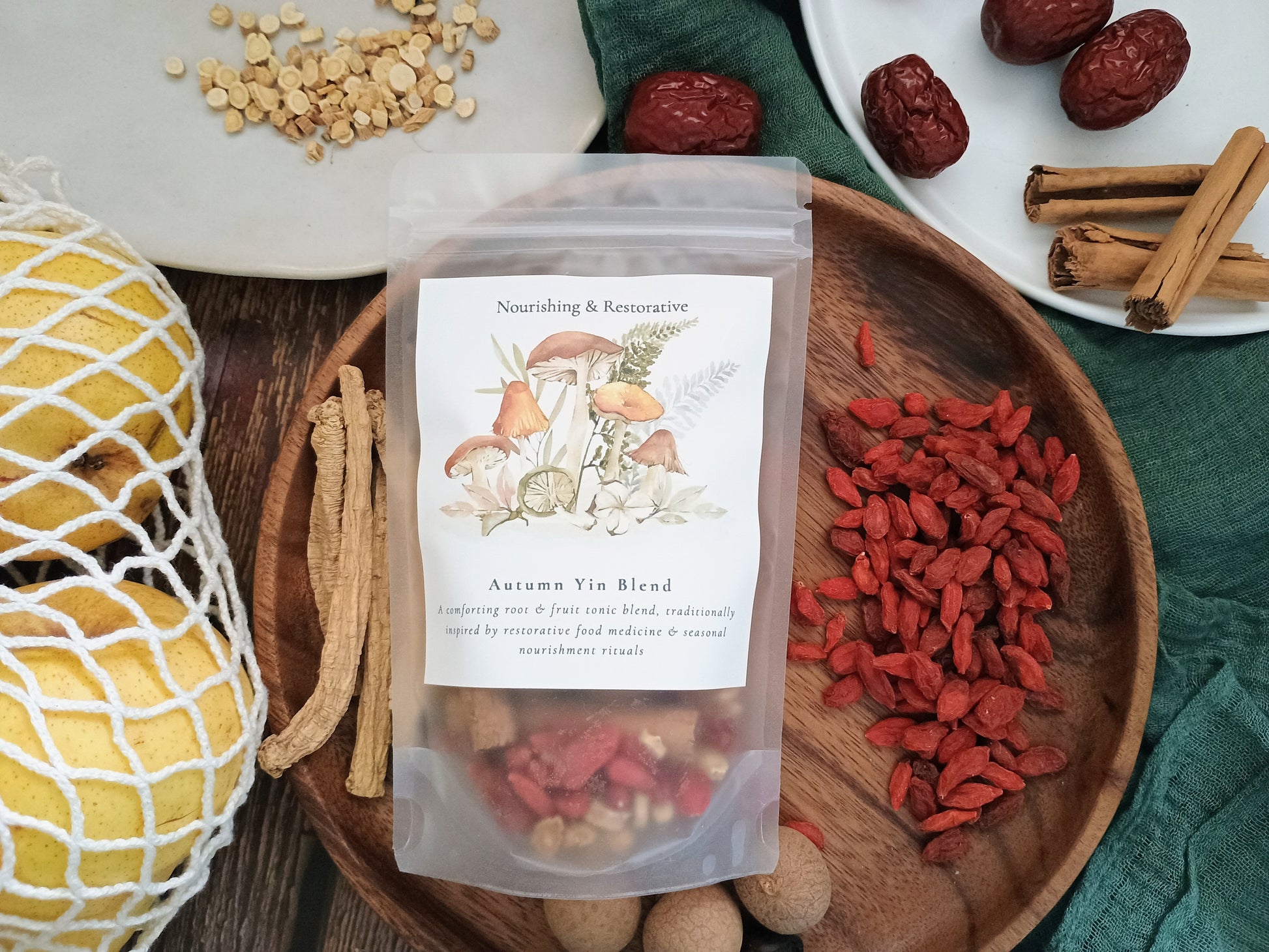 A stand-up pouch labeled 'Autumn Yin Blend' sits on a wooden tray surrounded by herbal ingredients including red dates, goji berries, cinnamon sticks, and a yellow gourd, with a green cloth background.