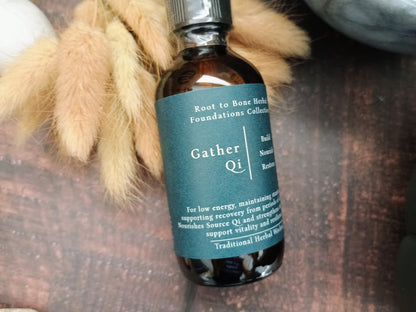 An amber glass bottle of Root to Bone Herbs 'Gather Qi' tincture, featuring a dark teal label, styled with dried bunny tails on a rustic wooden surface.