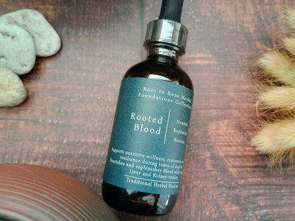 A dark amber glass dropper bottle labeled 'Rooted Blood' by Root to Bone Herbs, placed on a rustic wooden surface with decorative stones and dried grass.