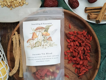A clear stand-up pouch labeled 'Autumn Yin Blend' with mushroom illustrations on the front, placed on a wooden tray alongside red goji berries, dried roots, cinnamon sticks, and other herbal ingredients.