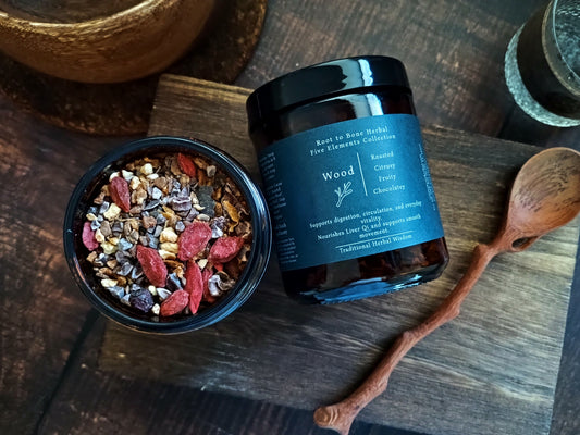 An open jar of Wood Root to Bone Herbal Five Elements Collection herbal tea blend sits next to its black-lidded jar on a rustic wooden board, with a wooden spoon and bowls in the background.