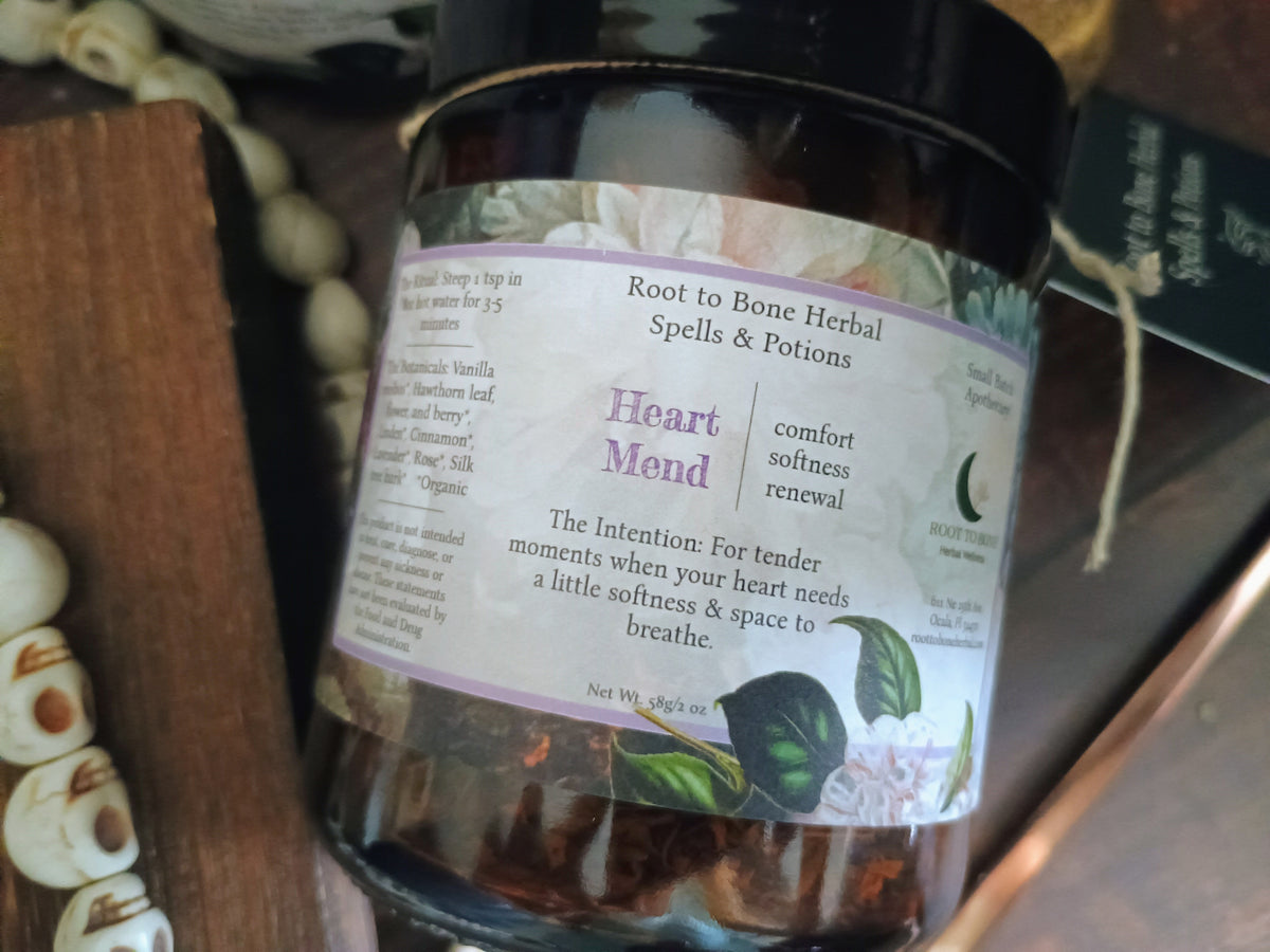 A glass jar of Root to Bone Herbal Spells & Potions, Heart Mend, filled with dried herbs, resting on a dark wooden surface next to white beads.
