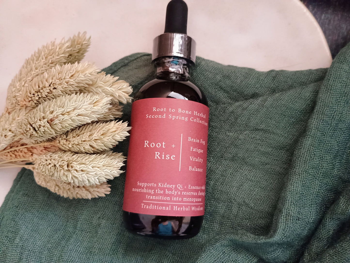 A dark amber glass dropper bottle with a black rubber bulb and silver collar, featuring a dusty rose pink label that reads 'Root to Bone Herbal Second Spring Collection Root + Rise'. The bottle is styled on a textured dark green knit blanket next to dried beige bunny tails on a neutral light background.