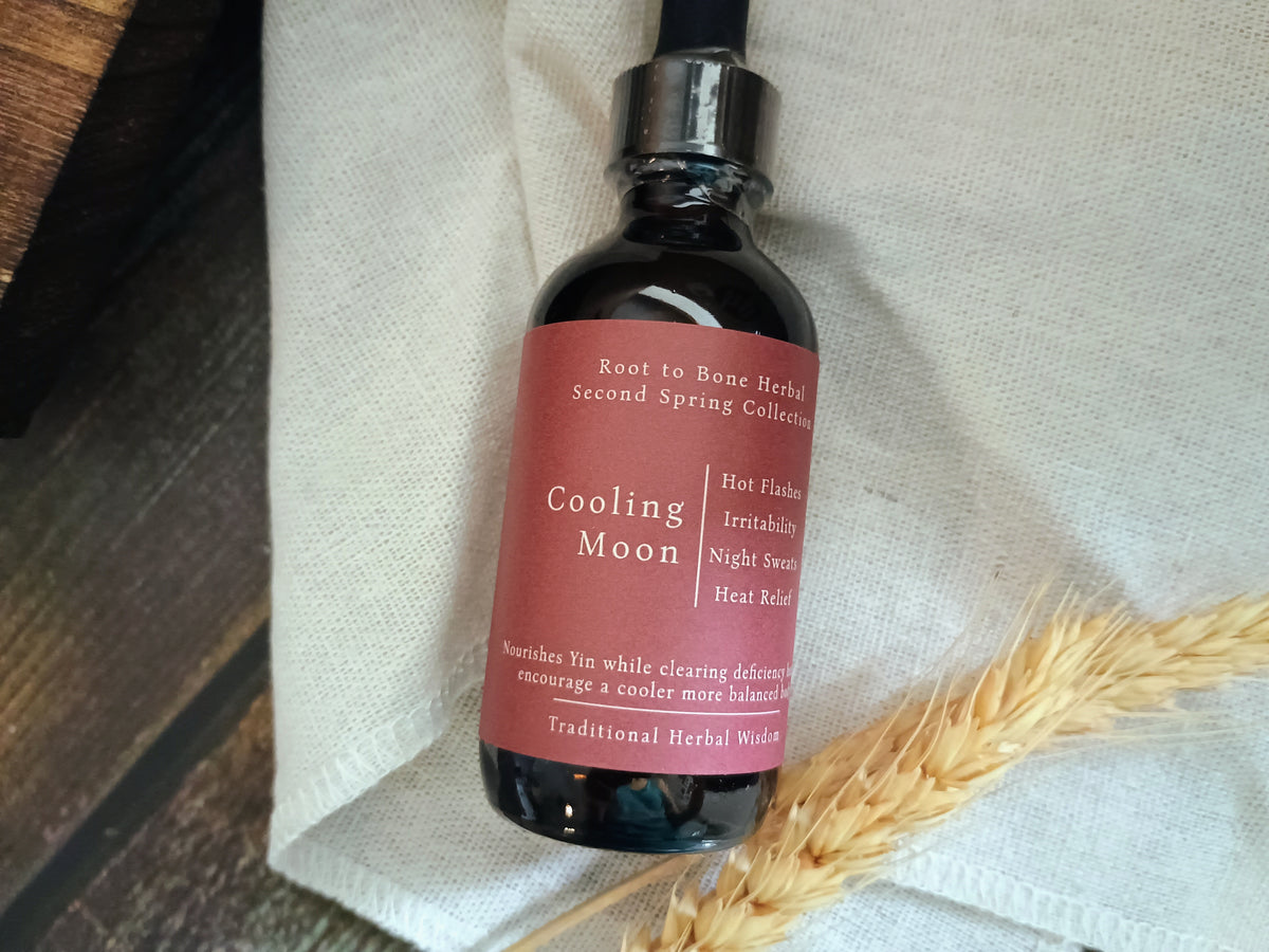 A dark amber glass dropper bottle labeled 'Cooling Moon' by Root to Bone Herbs, resting on a textured white linen cloth with dried wheat stalks.