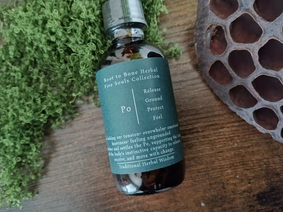 A small amber glass bottle with a dark teal label reading 'Root to Bone Herbal Five Souls Collection' and 'Po', styled on a wooden surface with green moss and a dried lotus pod.
