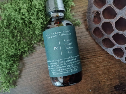 A small amber glass bottle with a dark teal label reading 'Root to Bone Herbal Five Souls Collection' and 'Po', styled on a wooden surface with green moss and a dried lotus pod.