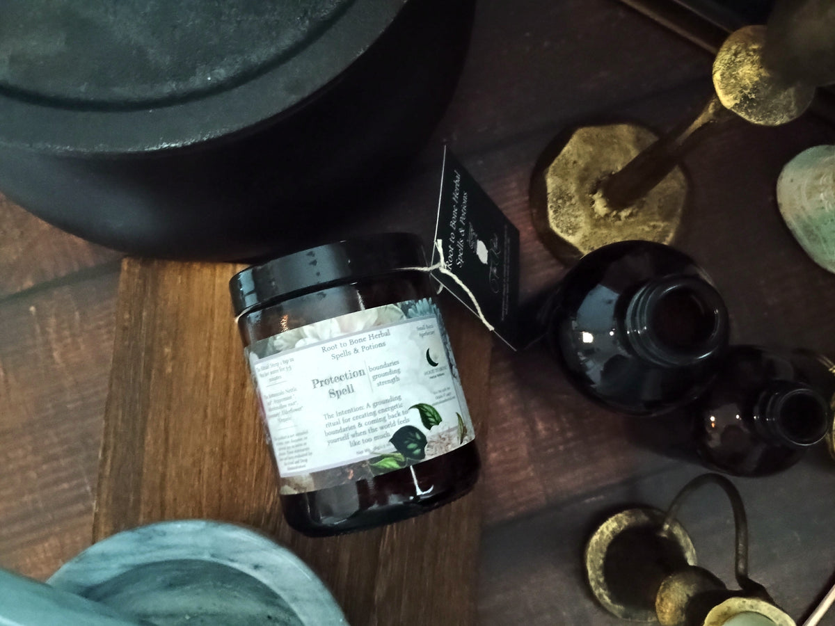 A jar of Protection Spell by Root to Bone Herbal, styled on a dark wooden surface with bohemian props including brass candle holders, a dark glass bottle, and a blue stone bowl.
