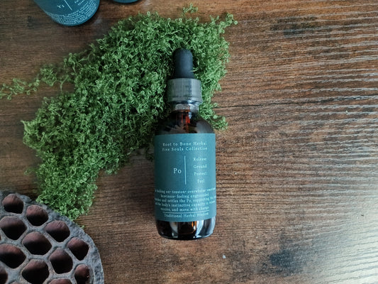 A dark amber glass dropper bottle with a muted teal label sits on a rustic wooden surface, surrounded by green moss and a dried lotus pod. The label reads 'Root to Bone Herbal' and 'Po'.