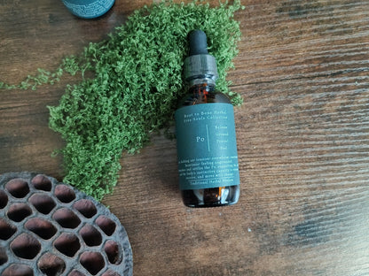 A small amber glass dropper bottle with a dark teal label sits on a rustic wooden surface, surrounded by green moss and a dark brown honeycomb-patterned dish.