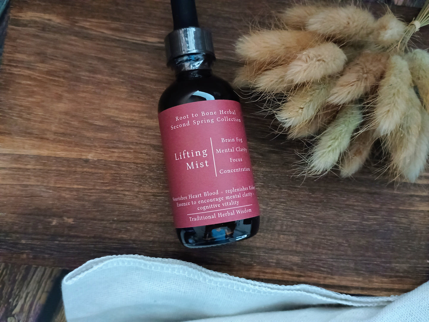 A bottle of Root to Bone Herbal Lifting Mist resting on a dark wooden surface next to dried bunny tail grass and a white cloth.