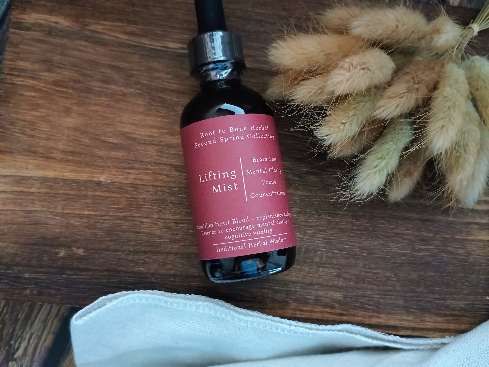 A bottle of Root to Bone Herbal Lifting Mist resting on a dark wooden surface next to dried bunny tail grass and a white cloth.