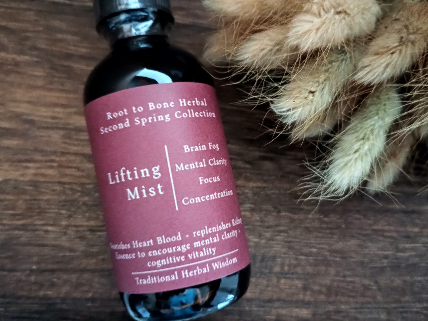 A dark glass bottle of Root to Bone Herbal Lifting Mist with a dusty rose label, resting on a dark wooden surface next to dried beige flowers.