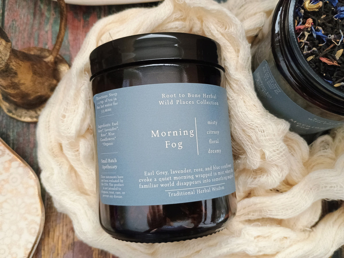 A dark amber glass jar candle with a black lid and a muted blue-gray label reading 'Morning Fog' from Root to Bone Herbal Wild Places Collection, styled on a rustic wooden surface with cream knit fabric and dried botanicals.
