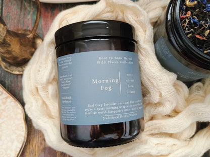 A dark amber glass jar candle with a black lid and a muted blue-gray label reading 'Morning Fog' from Root to Bone Herbal Wild Places Collection, styled on a rustic wooden surface with cream knit fabric and dried botanicals.