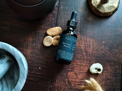 A dark amber glass dropper bottle labeled 'Moving Blood' by Root to bone herbal, styled on a dark wooden tabletop with small stones, a skull, and a mortar bowl nearby.