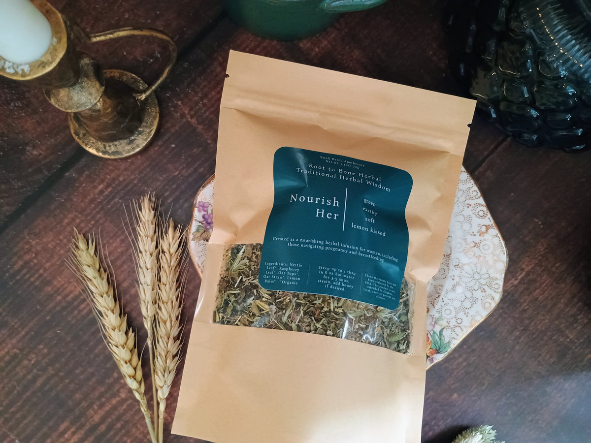 A kraft paper stand-up pouch labeled 'Nourish Her' containing loose herbal tea, styled on a dark wooden tabletop with wheat stalks, a candle holder, and decorative plates.