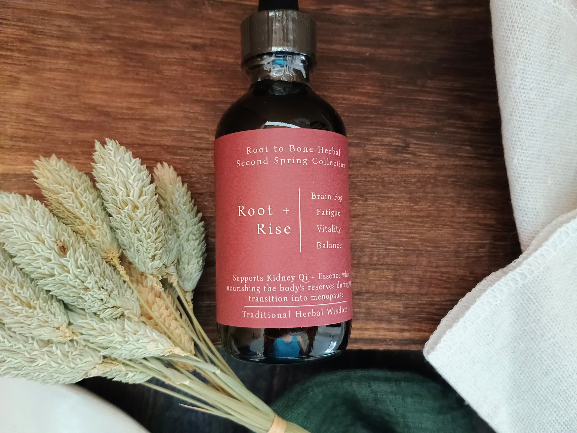 A dark amber glass bottle of Root to Bone Herbal Second Spring Collection Root + Rise tincture, featuring a dusty rose label, styled on a dark wood background with dried bunny tail grass and a white linen cloth.