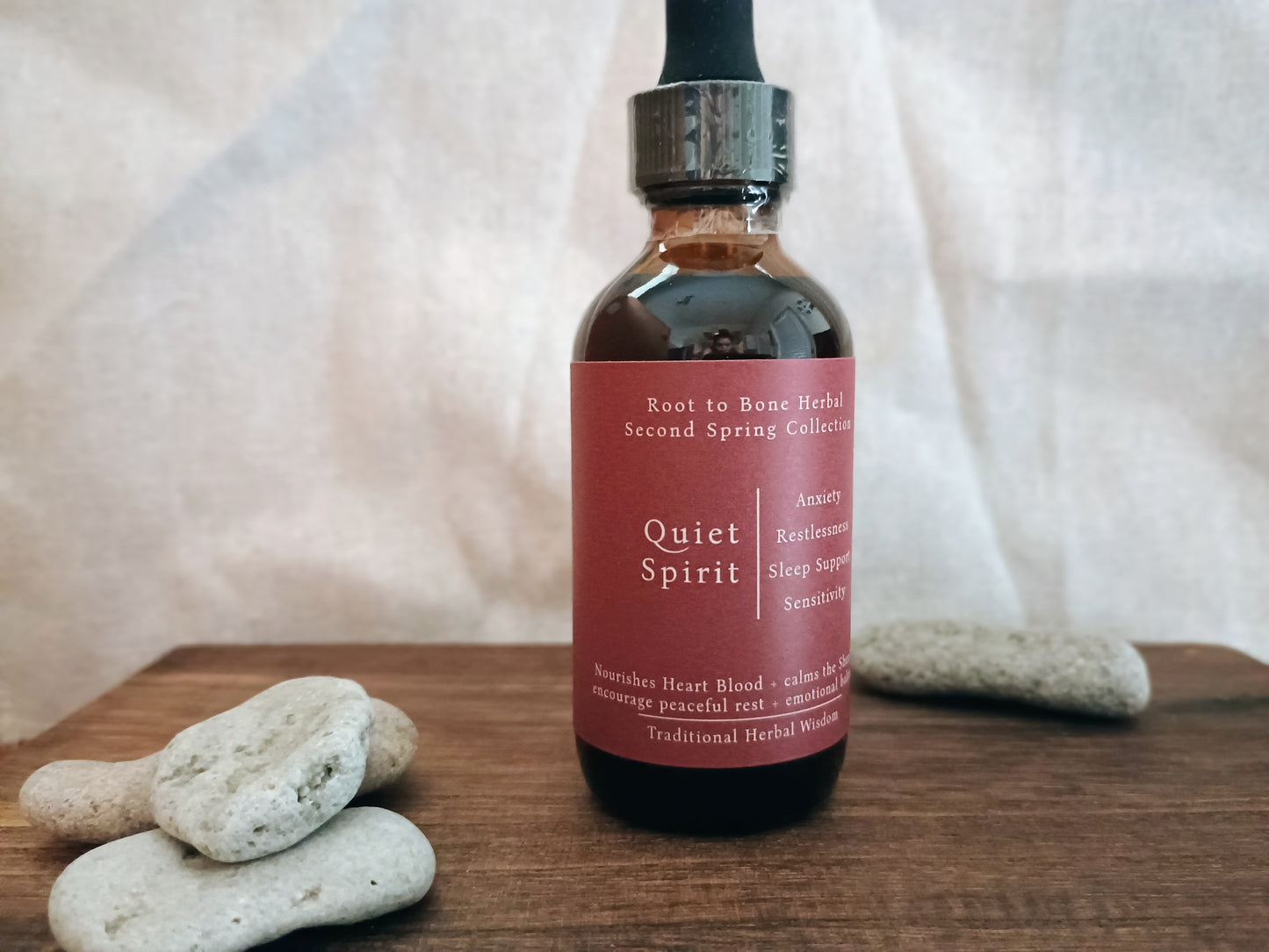 A dark amber glass dropper bottle labeled 'Quiet Spirit' by Root to Bone Herbal, set on a wooden surface with smooth stones against a draped fabric backdrop.