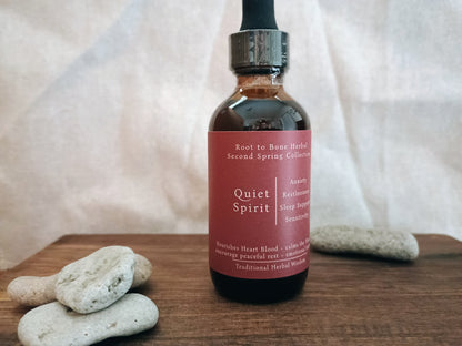 A dark amber glass dropper bottle labeled 'Quiet Spirit' by Root to Bone Herbal, set on a wooden surface with smooth stones against a draped fabric backdrop.