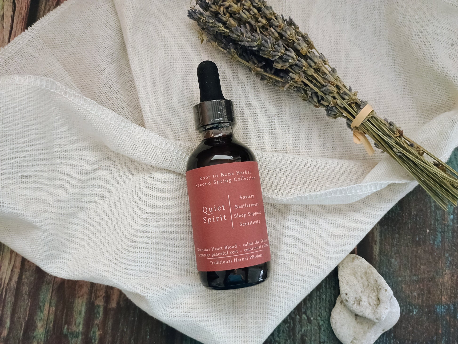 A small amber glass dropper bottle labeled 'Quiet Spirit' with a dusty rose label, placed on a cream linen cloth with dried lavender and small stones on a rustic wooden surface.