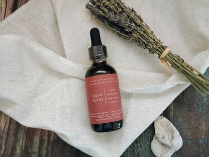 A small amber glass dropper bottle labeled 'Quiet Spirit' with a dusty rose label, placed on a cream linen cloth with dried lavender and small stones on a rustic wooden surface.