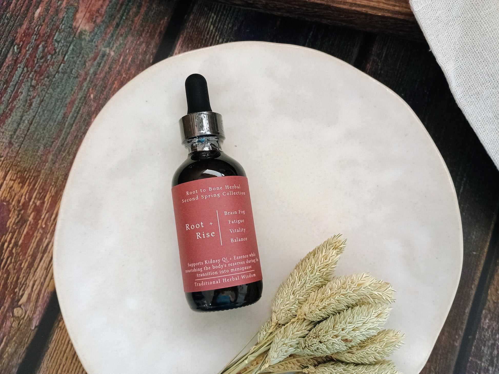 A small amber glass dropper bottle with a dusty rose label reading Root + Rise, placed on a round white plate with dried wheat stems on a rustic wooden surface.
